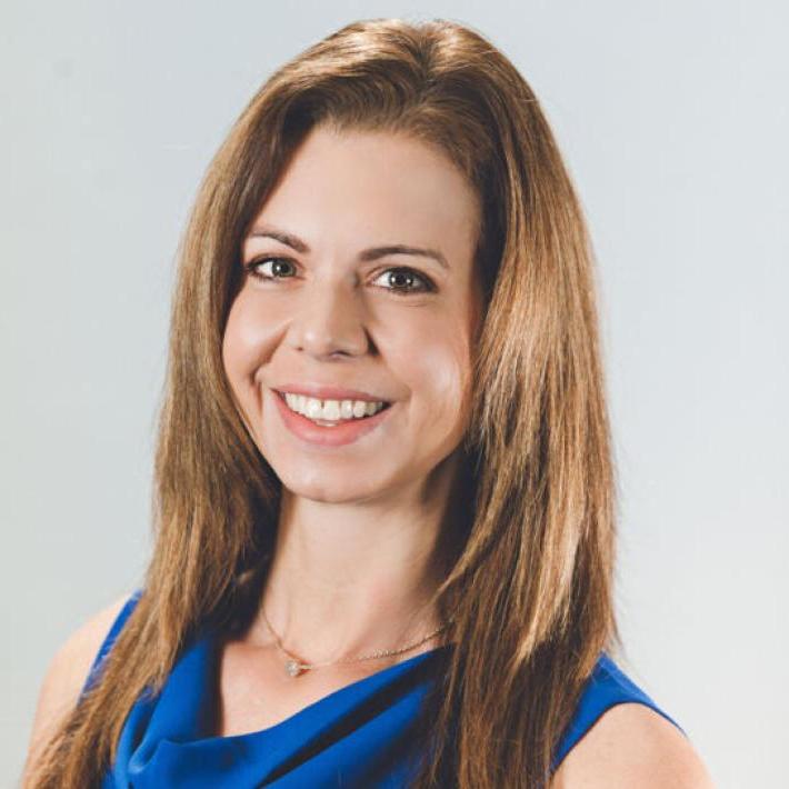 The image shows a woman with long, straight light brown hair wearing a sleeveless blue dress with a cowl neckline. She has a friendly expression with a broad smile and is facing slightly to her right. The background is a plain light gray. She has light skin and an approachable demeanor.